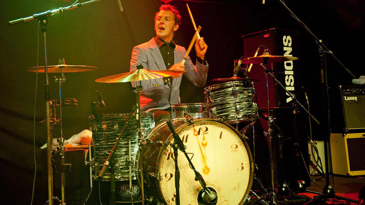 Man sieht den Schlagzeuger und sein Instrument auf dessen Bass-Drum eine große Uhr zu sehen ist.