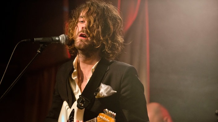Mann mit Locken, die ihm ins Gesicht fallen, singt in ein Mikrofon und spielt Gitarre