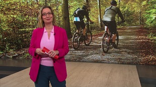  Yvonne Willicks steht im Servicezeit-Studio. Im Hintergrund sieht man zwei radfahrende Personen im Wald.