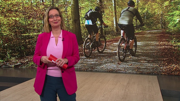  Yvonne Willicks steht im Servicezeit-Studio. Im Hintergrund sieht man zwei radfahrende Personen im Wald.