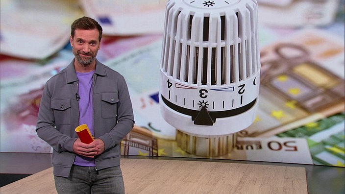 Daniel Aßmann steht im Servicezeit-Studio. Im Hintergrund sieht man die Nahaufnahme eines Heizungsreglers.