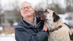 Ein Mann mit hellen Haaren und einer Brille umarmt einen großen beigefarbenen Hund 