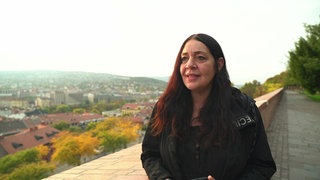 Eine Frau mit langen braunen Augen spricht, im Hintergrund ist ein Tal zu sehen 