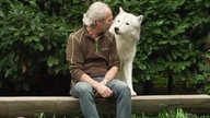 Ein Mann mit grauen Haare und braunem Oberteil sitzt gemeinsam mit einem weißen Wolf eng beieinander 