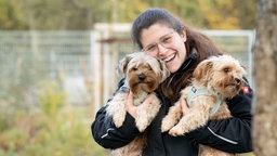 Eine dunkelhaarige Frau mit Brille trägt zwei beigefarbene Hunde auf dem Arm 