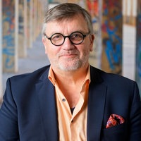 Hape Kerkeling schaut freundlich in die Kamera; er steht in einer leeren Fabrikhalle zwischen zwei Reihen von Betonsäulen, die mit bunten Graffiti bemalt wurden