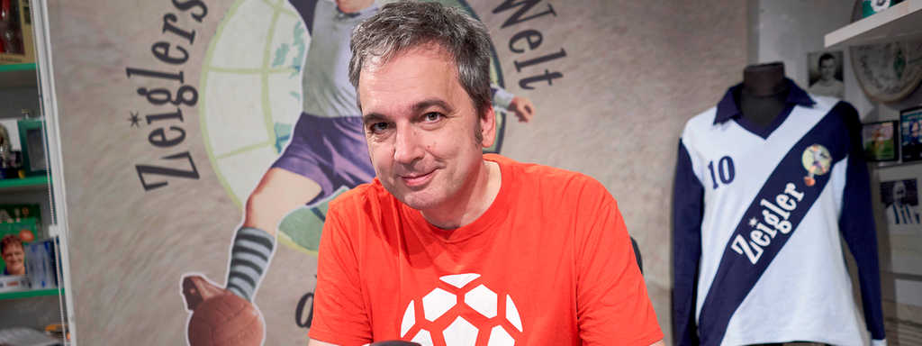 Arnd Zeigler sitzt in seinem Studio an seinem Schreibtisch und hält einen Fußball in den Händen