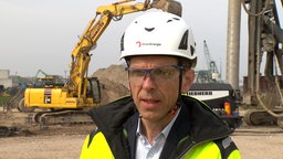 Ein Mann mit Helm und leuchtend gelber Jacke vor einer Baustelle