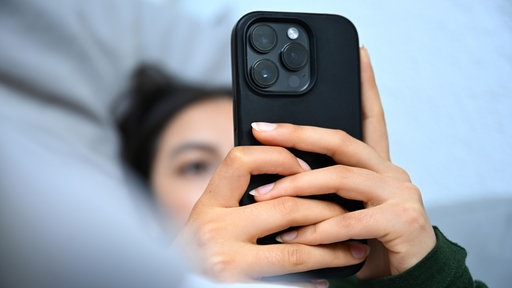 Eine junge Frau liegt mit ihrem Handy auf der Couch und blickt auf das Display
