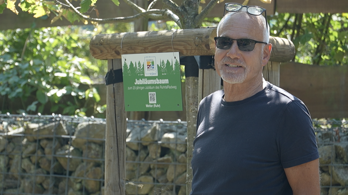 Peter Uphoff steht mit Sonnenbrille vor einer grünen Plakette mit Aufschrift "Jubiläumsbaum"