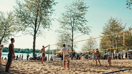 Personen spielen Volleyball auf einem Beachvolleyballfeld an einem See.