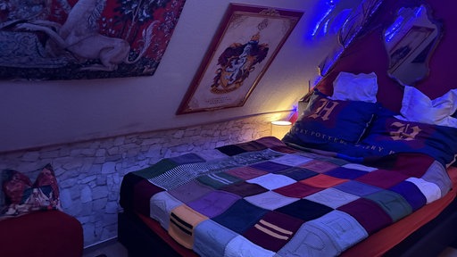 In der Mitte des Schlafzimmers steht ein Bett auf dem eine gestrickte Decke liegt. An der Wand hängt das Gryffindor-Wappen.