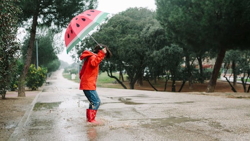 Ein Junge mit roter Regenjacke und roten Gummistiefel springt auf einer nassen Straße in eine Pfütze, braunes Wasser spritzt hoch, in seiner Hand hält er einen Regenschirm mit einem Wassermelonen-Muster