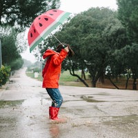 Ein Junge mit roter Regenjacke und roten Gummistiefel springt auf einer nassen Straße in eine Pfütze, braunes Wasser spritzt hoch, in seiner Hand hält er einen Regenschirm mit einem Wassermelonen-Muster