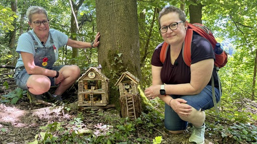 2 Frauen knien im Wald neben zwei aus Holz gebauten Wichtelhäuschen