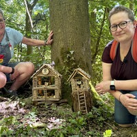 2 Frauen knien im Wald neben zwei aus Holz gebauten Wichtelhäuschen