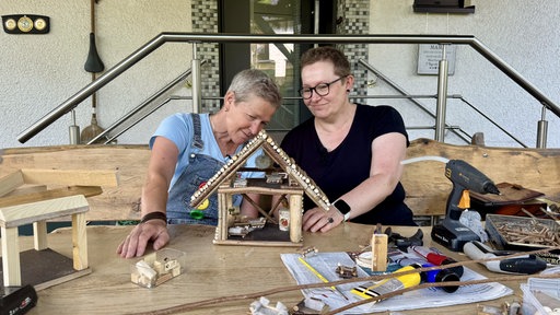 Zwei Frauen sitzen an einem großen Holztisch und schauen sich ein Wichtelhäuschen an