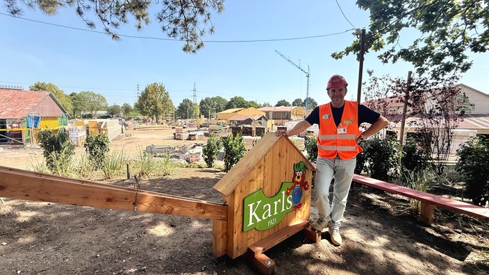 Auf dem Gelände der Baustelle von Karls Erlebnisdorf in Oberhausen steht Robert Dahl. Er stützt sich auf ein Schild mit der Aufschrift "Karls", dabei trägt er einen Bauhelm und eine Warnweste.