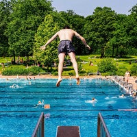 Junge springt vornüber von einem 3-Meter-Brett, steht in der Luft, unter ihm der Pool mit einigen Menschen im Pool und am Beckenrand, in naher Ferne Liegewiese und Wald 