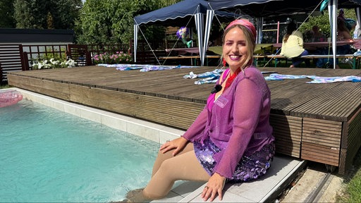 Veranstalterin Janina Gierke sitzt in einem pinken Outfit am Pool und hält ihre Beine ins Wasser. Sie lächelt in die Kamera.