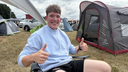 Ein Mann sitzt im Campingstuhl auf einem Campingplatz und hält beide Daumen hoch.