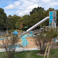 Ein Spielplatz mit großen blauen und beigen Spielgeräten umgeben von Bäumen