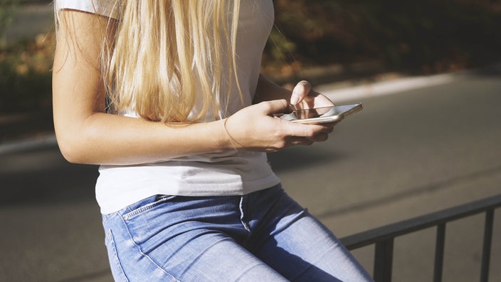 Ein junges Mädchen sitzt auf einem Geländer und blickt aufs Handy, das Gesicht ist nicht zu erkennen