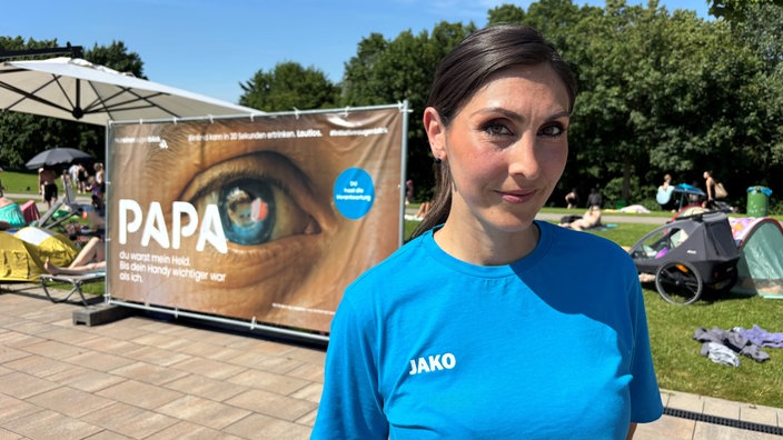 Eine Frau mit blauem Oberteil steht vor einem Schild. Auf dem Schild ist ein Auge drauf und im Auge ist ein Kind zu sehen was im Wasser ist.