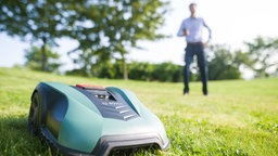 Ein Mähroboter auf einer Wiese, im Hintergrund steht ein Mann
