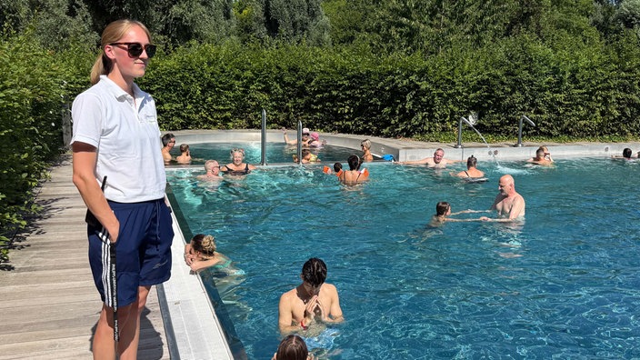 Eine Mitarbeiterin des Schwimmbads steht am Beckenrand und schaut auf viele Badegäste im Wasser.