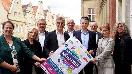 Neun Personen, darunter Bürgermeister Tilman Fuchs, halten ein Plakat des NRW-Tags 2026 auf dem Balkon des Münsteraner Rathauses hoch.