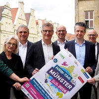Neun Personen, darunter Bürgermeister Tilman Fuchs, halten ein Plakat des NRW-Tags 2026 auf dem Balkon des Münsteraner Rathauses hoch.