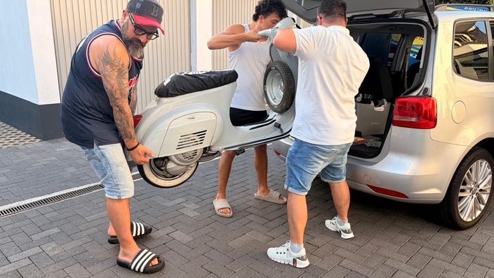 Drei Männer laden einen Vespa-Roller auf die Ladefläche eines Autos
