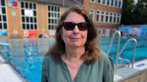 Portait von Jessica Mense, eine Frau mit langen dunklen Haaren und Sonnenbrille, im Hintergrund ist ein Schwimmbecken zu sehen 