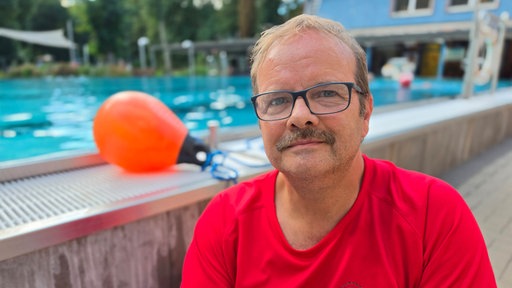 Portait von Georg Schwarzkopf, er trägt ein rotes T-Shirt, im Hintergrund ist ein Schwimmbecken zu sehen