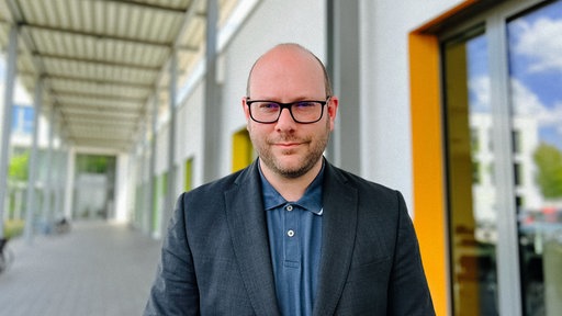 Manuel Heinemann steht in einem Gang vor einem Haus und schaut in Richtung Kamera