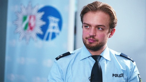 Hendrik Heyer, ein junger blonder Mann mit kurzen Haaren im Mittelscheitel und Vollbart, sitzt im blauen Hemd vor einer hellen Wand und schaut nach links aus dem Bild.