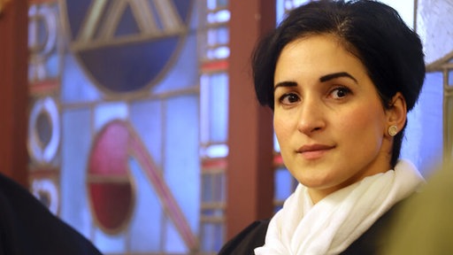 Gülkiz Yazir schaut entlang der Kamera. Sie hat dunkle Haare und trägt ein weißes Halstuch.