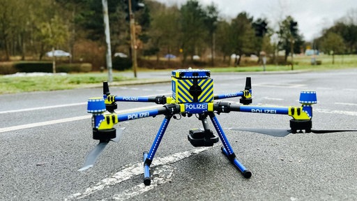 Auf der Straße steht eine Drohne. Sie ist blau und gelb mit der Aufschrift "Polizei".