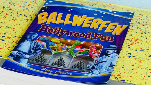 Der Flyer zur Wurfbude "Hollywood Fun" liegt auf einer Oberfläche. Darauf befindet sich ein Foto mit Türmen aus Dosen und Kuscheltieren darüber.