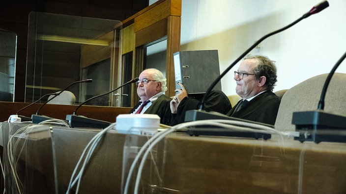 Drei Männer sitzen an einem Tisch mit Mikrofonen, einer der Männer hält sich einen Aktenorder vor das Gesicht.