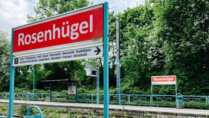 Das Haltestellenschild Rosenhügel in Köln. Im Hintergrund die Straßenbahnschienen und Bäume.