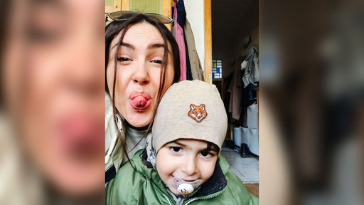 Derya trägt einen Sonnenbrille auf dem Kopf und ihr Sohn Kian eine graue Mütze mit einem Fuchs drauf. Die beiden machen ein Selfie. Derya streckt ihre Zunge heraus und Kian lächelt mit Schnuller im Mund.