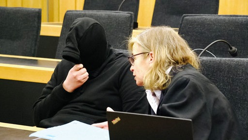 Ein Mann sitzt mit einer schwarzer Kapuze vor dem Gesicht in einem Gerichtssaal. Seine Rechtsanwältin spricht zu ihm.