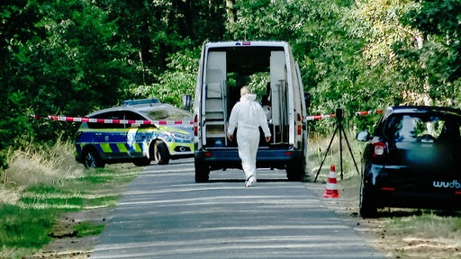 Ein abgesperrter Straßenabschnitt, an dem eine Person in weißem Schutzanzug zu einem geöffneten Transporter geht. Im Hintergrund ist ein Polizeifahrzeug und Absperrband zu sehen.