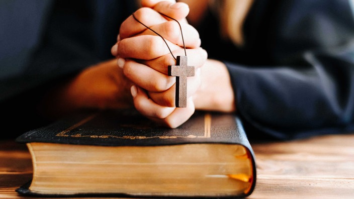 Hände einer Frau sind über einem schwarzen Buch gefaltet, über den Händen hängt ein Holzkreuz an einer Kette