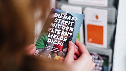 Die Kamera schaut einer Person über die Schulter. Sie hält einen Flyer mit der Aufschrift "Du hast Streit mit den Eltern? Melde Dich!" in der Hand.