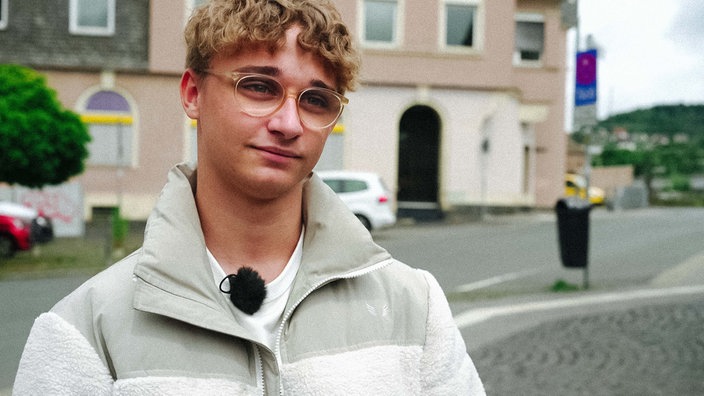Ein blonder Junge mit Brille schaut an der Kamera vorbei, im Hintergrund sind Häuser zu sehen.