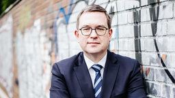Markus Hartmann, Brille, kurze, blonde Haare, blaues Jaquette, weißes Hemd und blau-gestreifte Krawatte, Blick nach vorne, hinter ihm eine Mauer mit Graffiti 