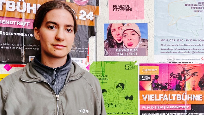 Lilly, eine junge Frau, mit dunklen Haaren, steht vor einer Wand, an der mehrere Plakate angebracht sind. Darunter ein Foto von Derya Seyhun und ihrem Sohn.